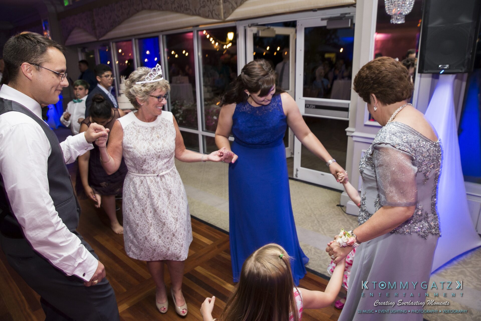 Jamie-and-Mark-Low-Res-Watermark-758 - TOMMY GATZ ENTERTAINMENT Group of people, including a child, dancing at a formal event. Women in dresses and men in suits hold hands in a circle on a wooden floor.