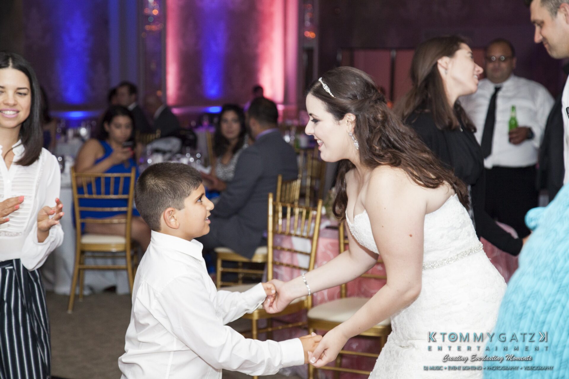Jamie-and-Mark-Low-Res-Watermark-762 - TOMMY GATZ ENTERTAINMENT A bride in a white dress dances with a young boy in a white shirt at a wedding reception. Guests are seated in the background.