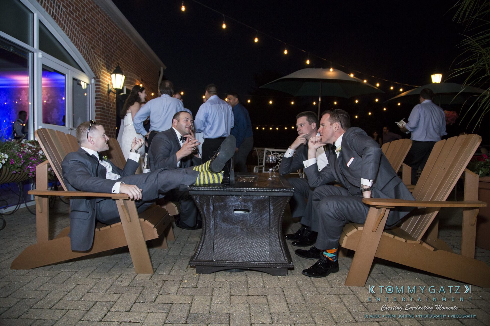 Jamie-and-Mark-Low-Res-Watermark-780 - TOMMY GATZ ENTERTAINMENT Four men in suits sit on patio chairs, engaged in conversation. One has feet up, displaying striped socks. Evening setting with decorative lights overhead.