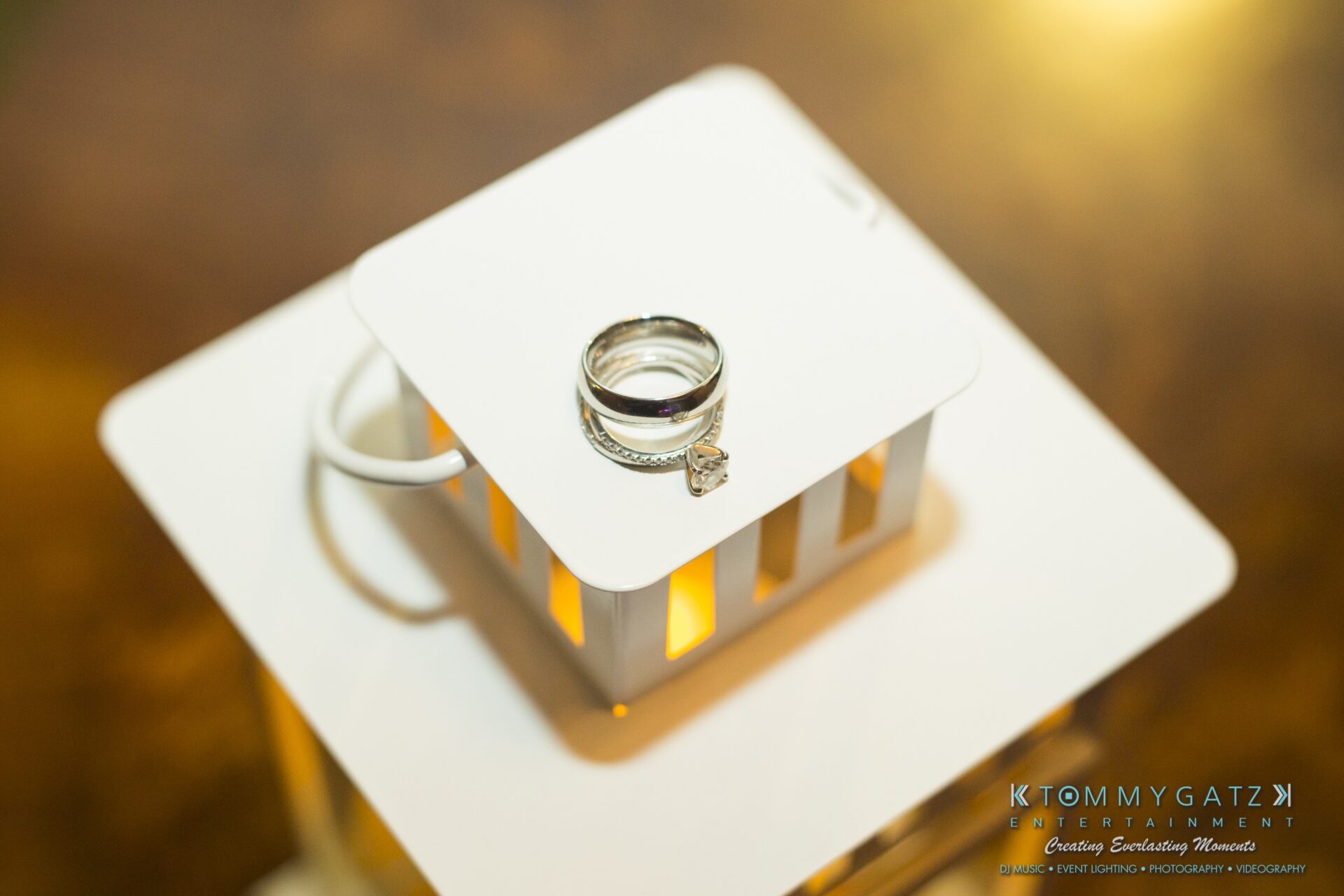 Jamie-and-Mark-Low-Res-Watermark-782 - TOMMY GATZ ENTERTAINMENT A set of wedding and engagement rings placed on a small white lantern with a warm light glow.