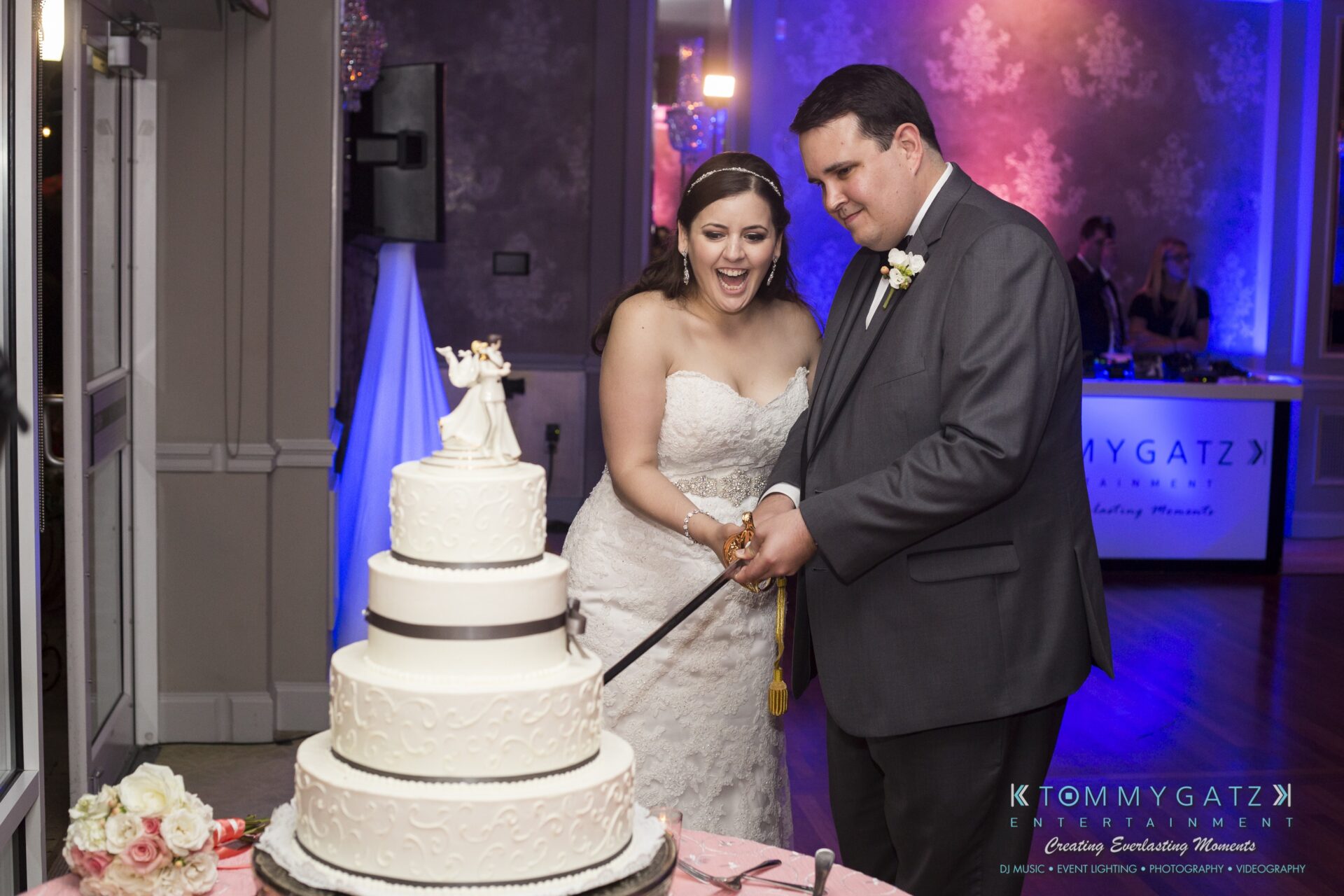 Jamie-and-Mark-Low-Res-Watermark-784 - TOMMY GATZ ENTERTAINMENT A bride and groom cut a multi-tiered white wedding cake together in a decorated venue.