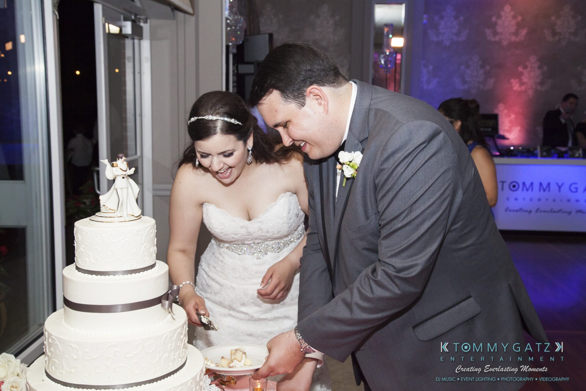 Jamie-and-Mark-Low-Res-Watermark-786 - TOMMY GATZ ENTERTAINMENT A bride and groom are cutting a tiered white wedding cake together.