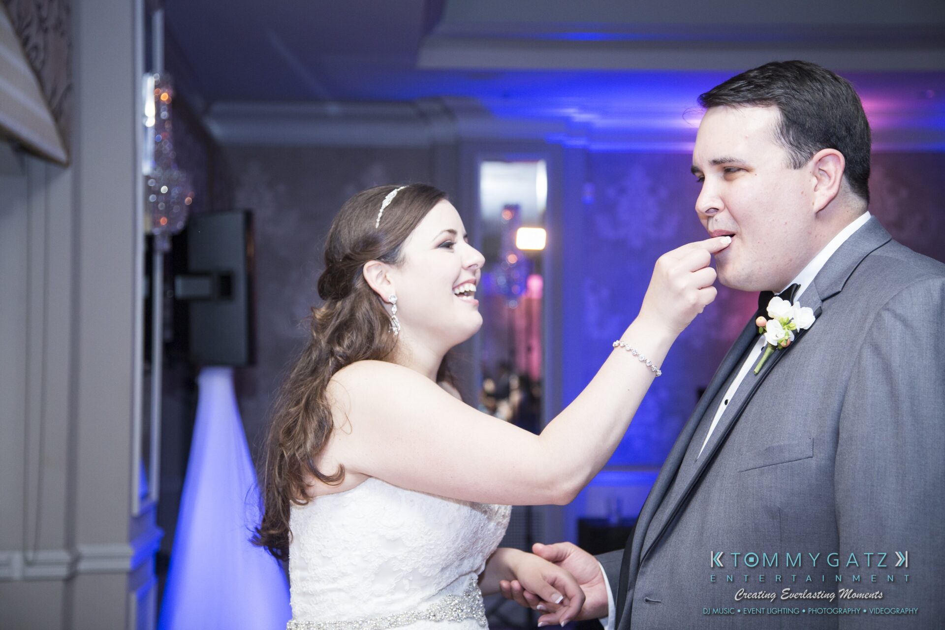 Jamie-and-Mark-Low-Res-Watermark-787 - TOMMY GATZ ENTERTAINMENT A bride playfully feeds cake to a groom in a decorated room with dim lighting.