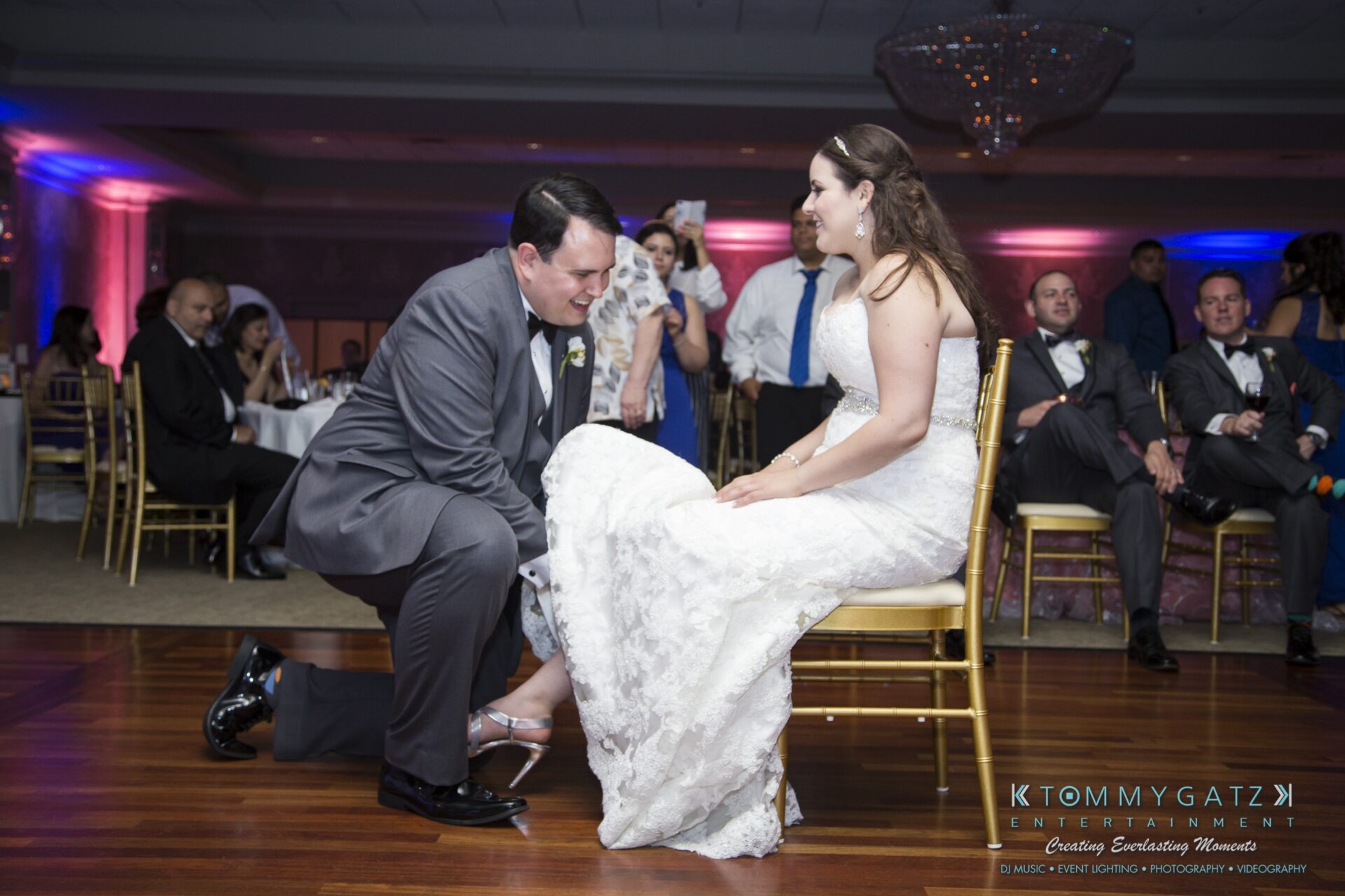 Jamie-and-Mark-Low-Res-Watermark-788 - TOMMY GATZ ENTERTAINMENT A groom kneels and removes a garter from the bride's leg at a wedding reception while guests look on.