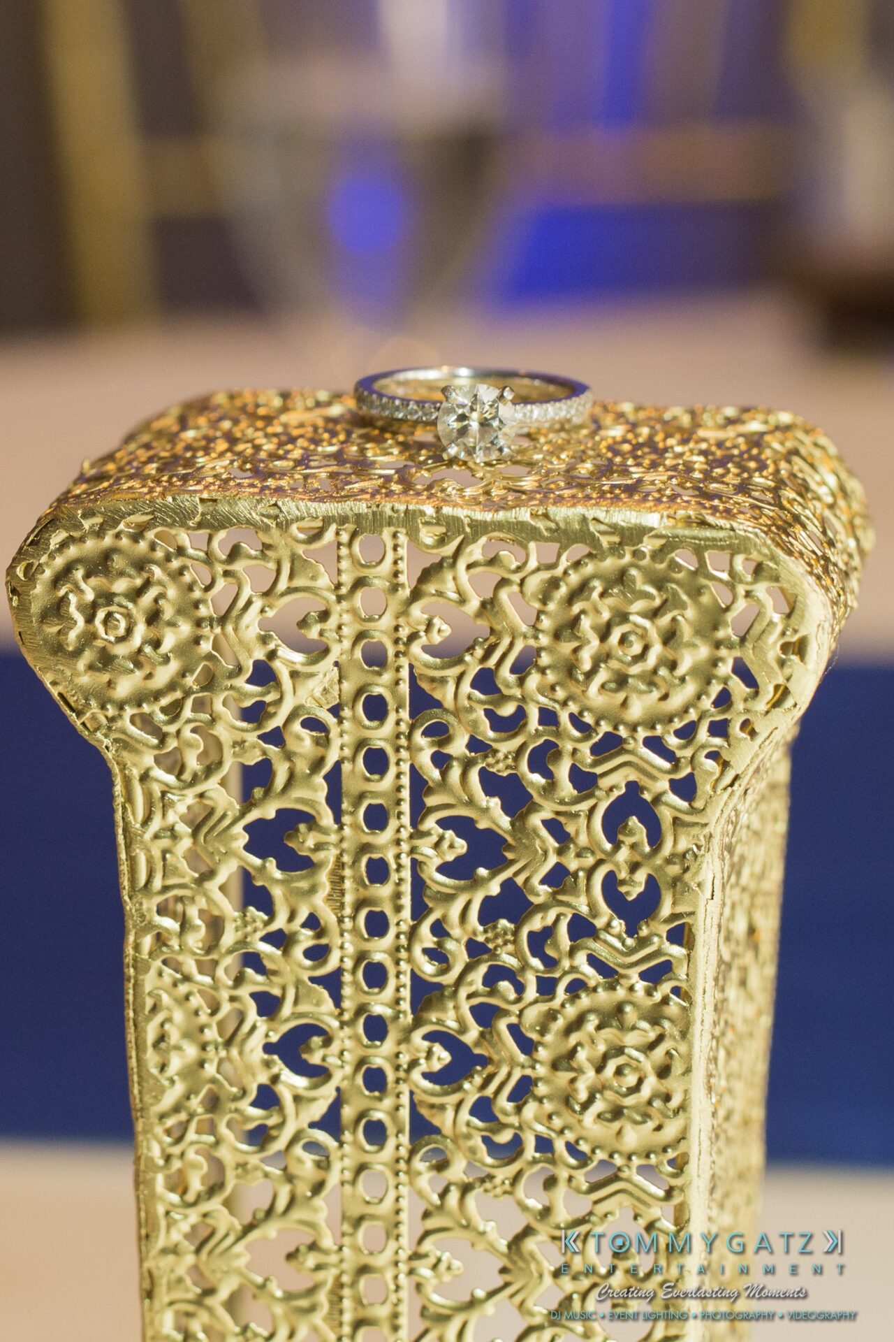 Jamie-and-Mark-Low-Res-Watermark-791 - TOMMY GATZ ENTERTAINMENT A silver ring with a diamond on an ornate, gold-colored textured surface.