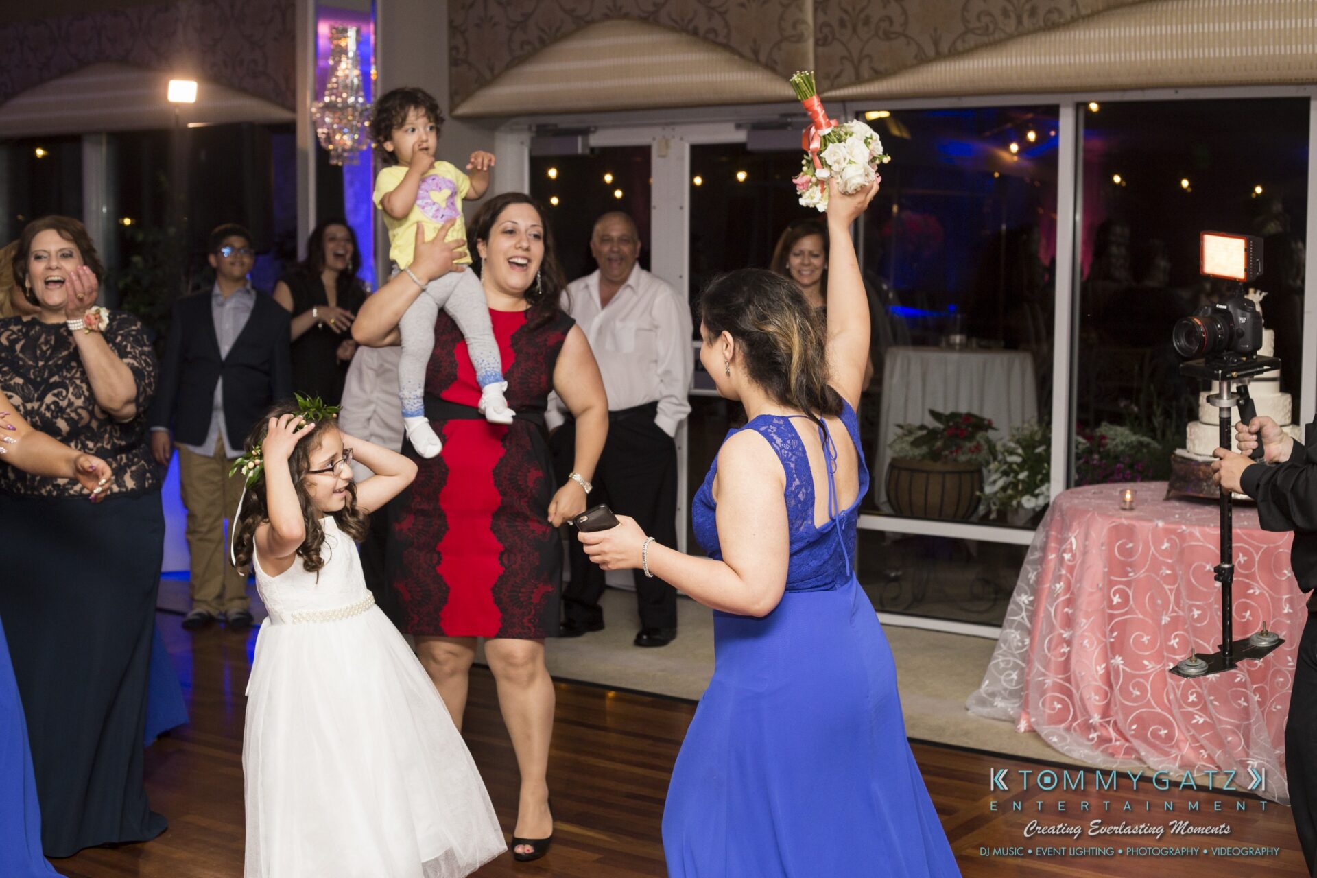 Jamie-and-Mark-Low-Res-Watermark-796 - TOMMY GATZ ENTERTAINMENT People dancing at a celebration, with a woman in a blue dress holding a bouquet. A child is on a woman's shoulders, and guests are smiling and watching the event.
