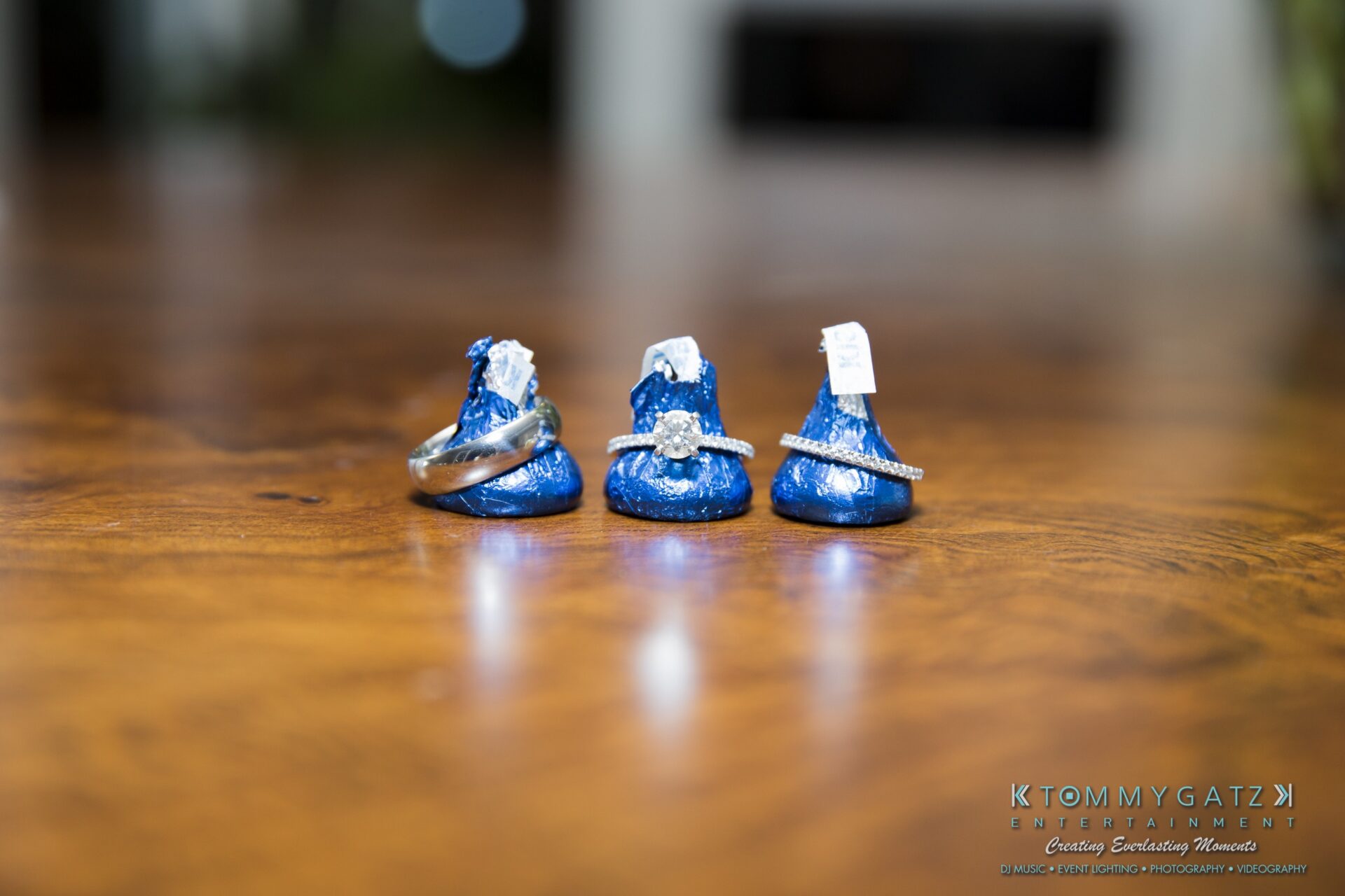 Jamie-and-Mark-Low-Res-Watermark-802 - TOMMY GATZ ENTERTAINMENT Three Hershey's Kisses hold two wedding bands and an engagement ring, placed on a wooden surface.