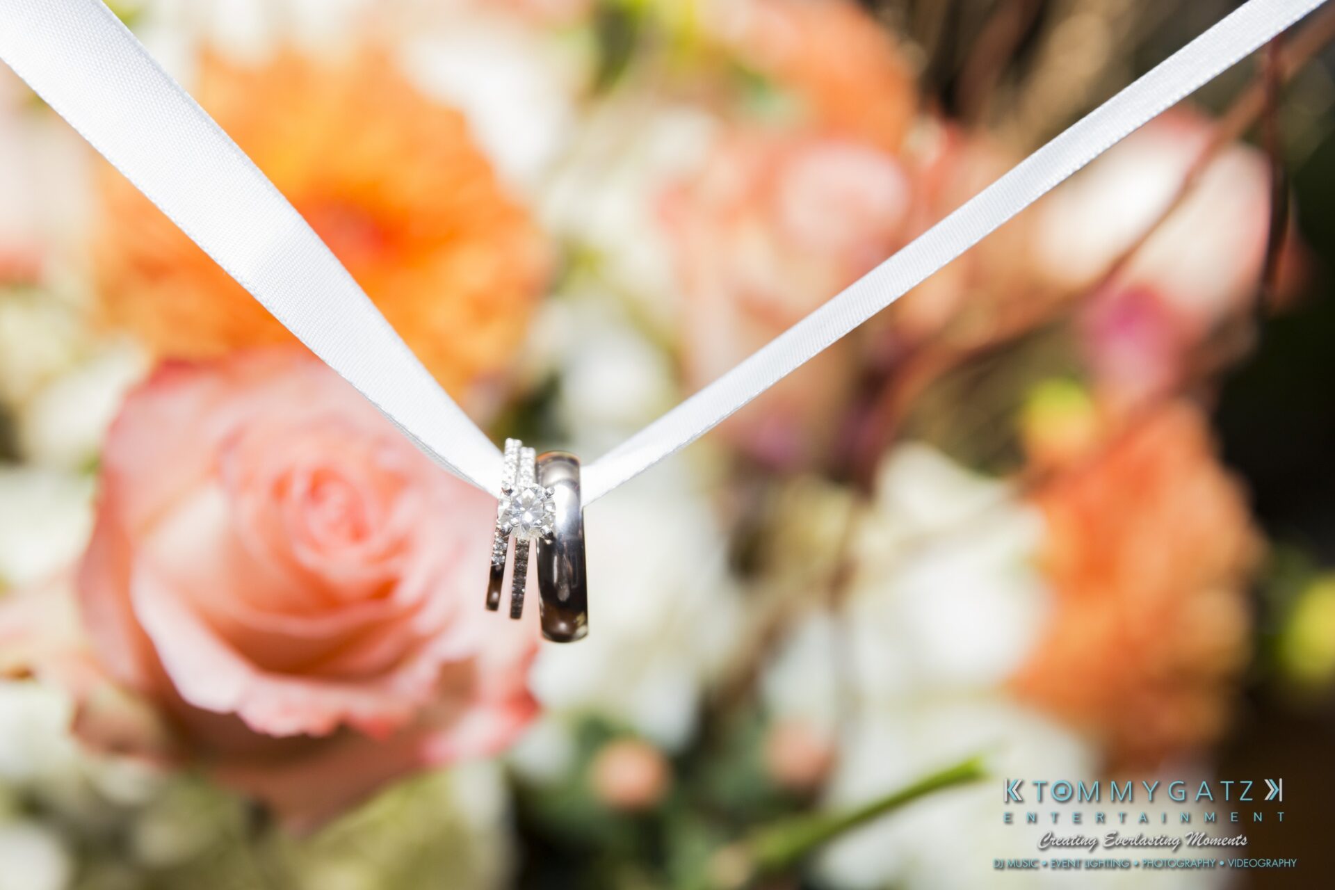 Jamie-and-Mark-Low-Res-Watermark-804 - TOMMY GATZ ENTERTAINMENT Close-up of wedding rings tied with a white ribbon against a blurred background of pink and orange flowers.