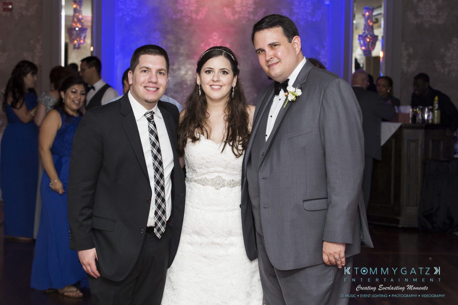 Jamie-and-Mark-Low-Res-Watermark-829 - TOMMY GATZ ENTERTAINMENT A bride stands between two men in suits at an indoor event, with guests and a DJ booth in the background.