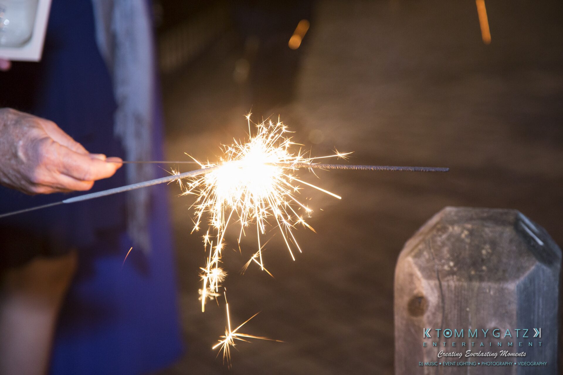 Jamie-and-Mark-Low-Res-Watermark-834 - TOMMY GATZ ENTERTAINMENT A person holding a lit sparkler in one hand with a blurred background.