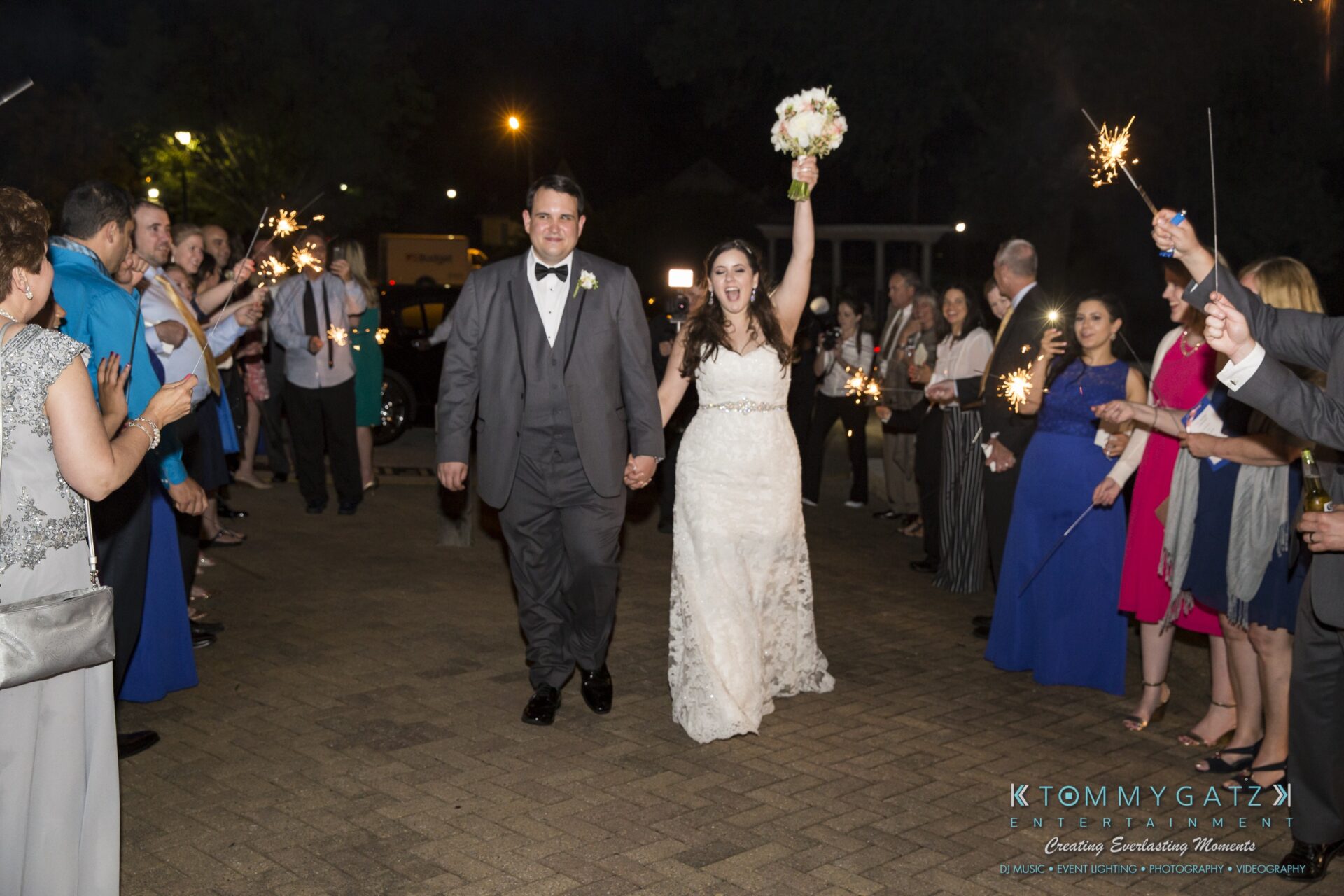 Jamie-and-Mark-Low-Res-Watermark-837 - TOMMY GATZ ENTERTAINMENT A bride and groom, holding hands, walk through a crowd of people holding sparklers. The bride is raising a bouquet in celebration.