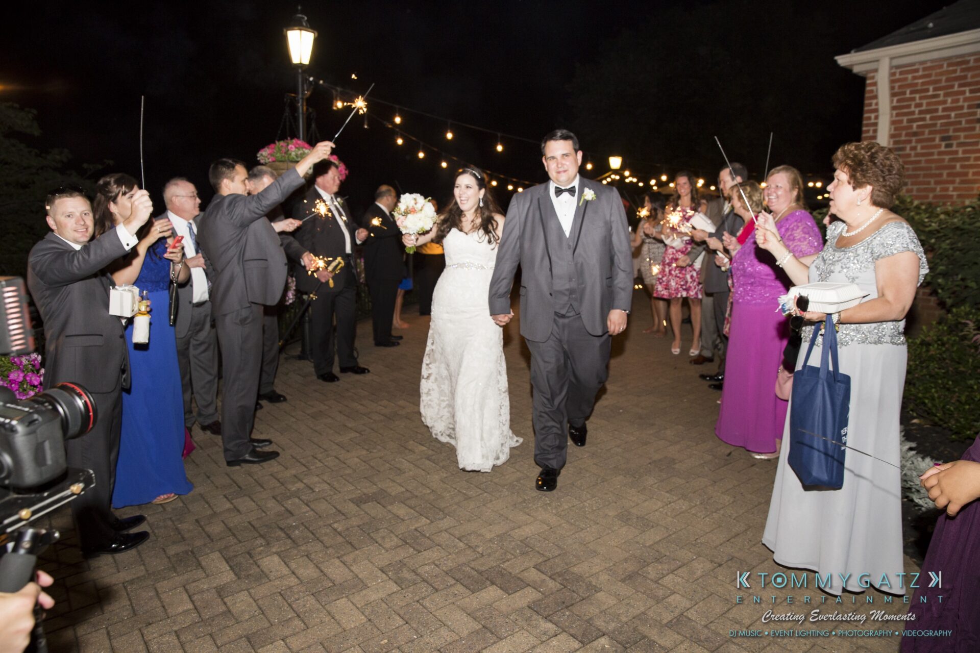 Jamie-and-Mark-Low-Res-Watermark-838 - TOMMY GATZ ENTERTAINMENT A bride and groom walk down a brick path under string lights, surrounded by people cheering and holding sparklers.