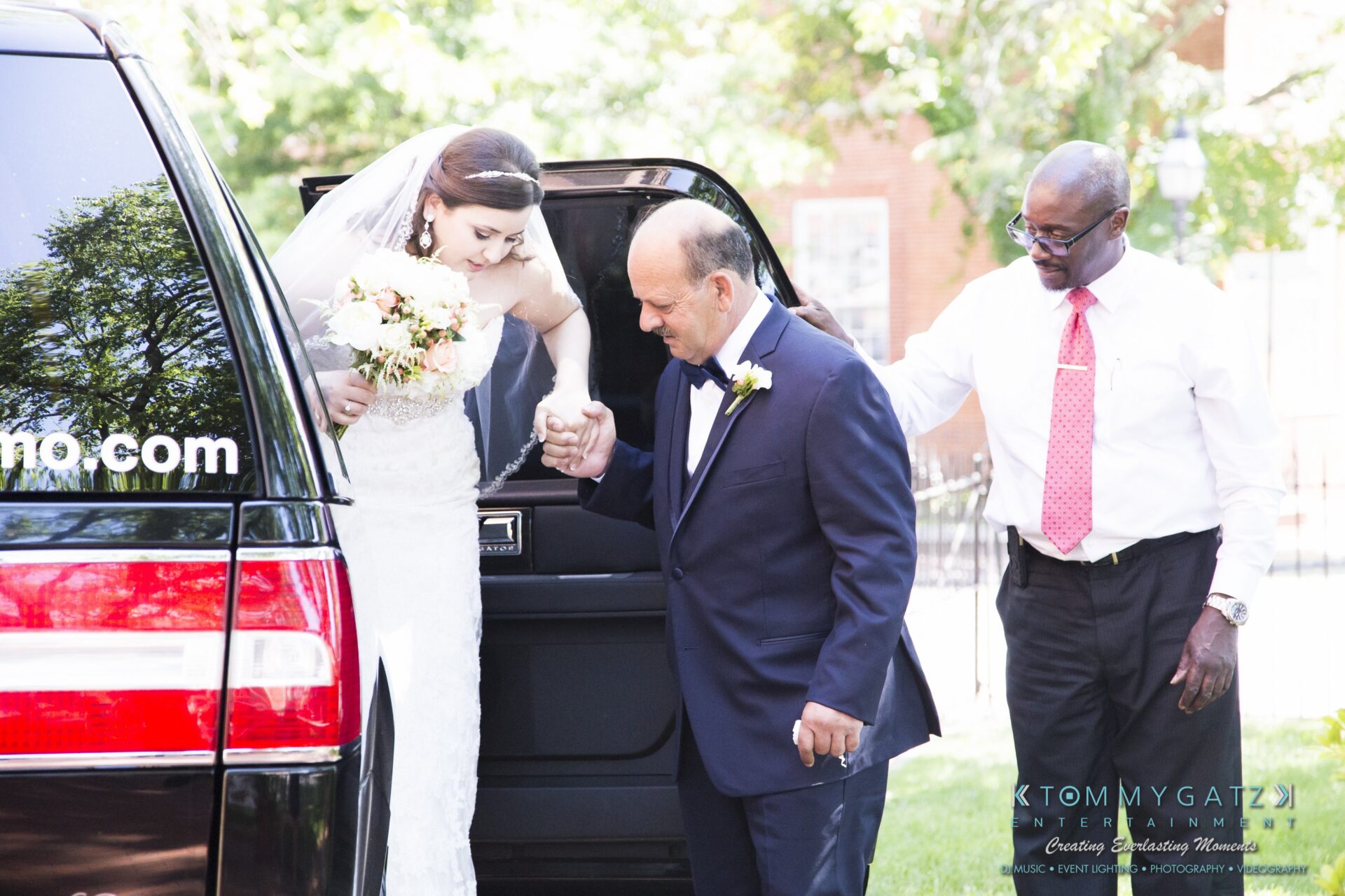 Jamie-and-Mark-Low-Res-Watermark-88 - TOMMY GATZ ENTERTAINMENT A bride steps out of a vehicle assisted by an older man in a suit, while another man stands nearby. The bride holds a bouquet and is wearing a veil.