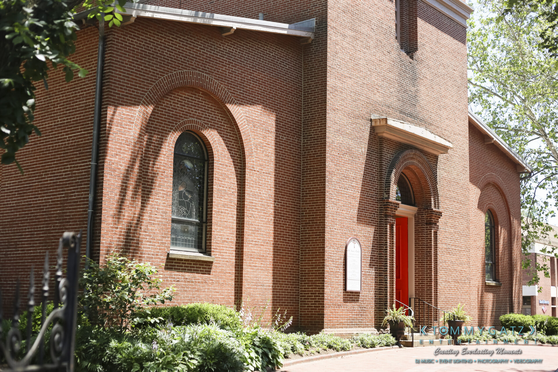 Jamie-and-Mark-Low-Res-Watermark-9 - TOMMY GATZ ENTERTAINMENT A red brick church with arched windows and a red door, surrounded by greenery and a wrought iron fence.