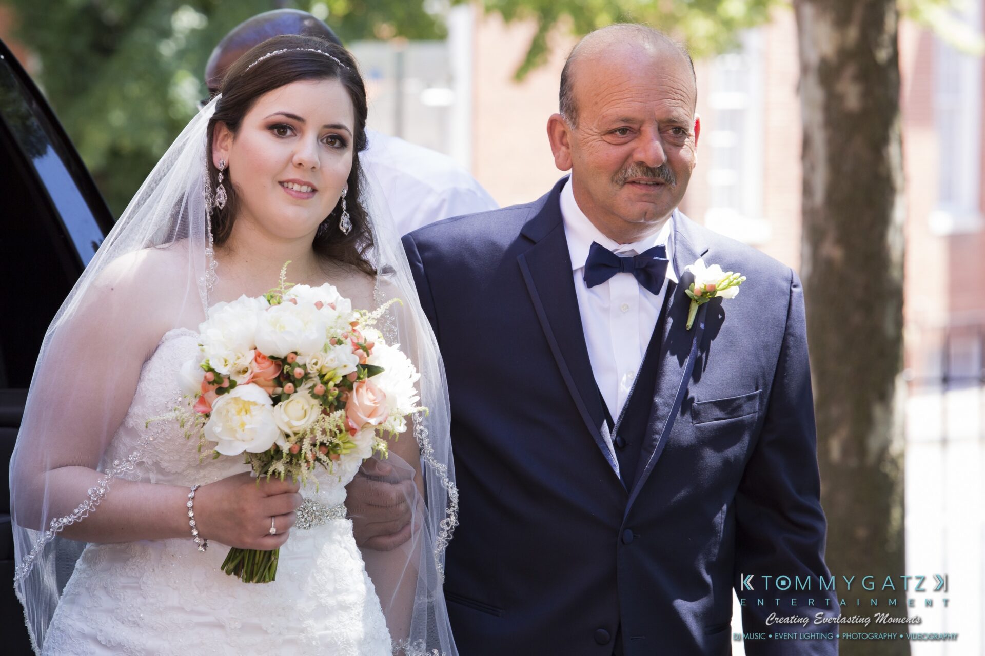 Jamie-and-Mark-Low-Res-Watermark-95 - TOMMY GATZ ENTERTAINMENT A bride in a white gown holds a bouquet, standing next to a man in a suit and bow tie. They are outdoors with a tree in the background.