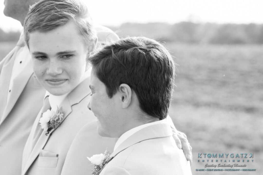 Jennifer-Rudi-Highlights-Low-res-22 - TOMMY GATZ ENTERTAINMENT Two young men in formal attire stand outdoors, exchanging a smile. They are wearing boutonnieres on their suits. The background is a blurred natural setting.