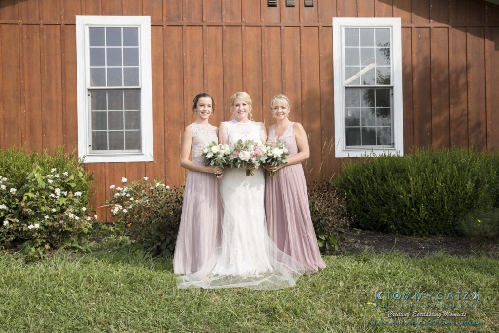 Jennifer-Rudi-Highlights-Low-res-36 - TOMMY GATZ ENTERTAINMENT Three women in formal dresses stand holding bouquets in front of a wooden building with two windows.