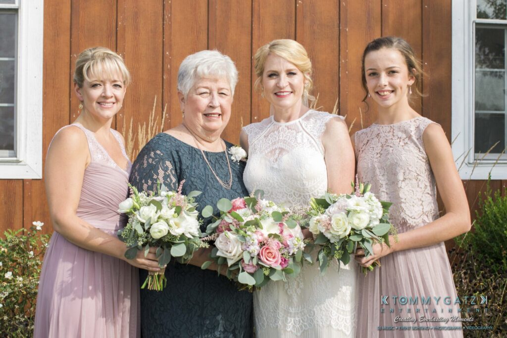 Jennifer-Rudi-Highlights-Low-res-40 - TOMMY GATZ ENTERTAINMENT Four women stand smiling, each holding bouquets, in front of a wooden wall. They are dressed in formal attire, and appear to be at a wedding.