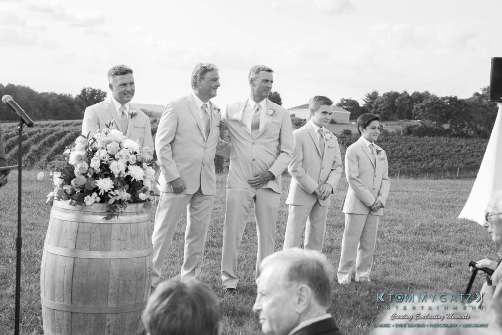 Jennifer-Rudi-Highlights-Low-res-8 - TOMMY GATZ ENTERTAINMENT A group of men in suits stand outside at a wedding ceremony near a wine barrel with flowers.
