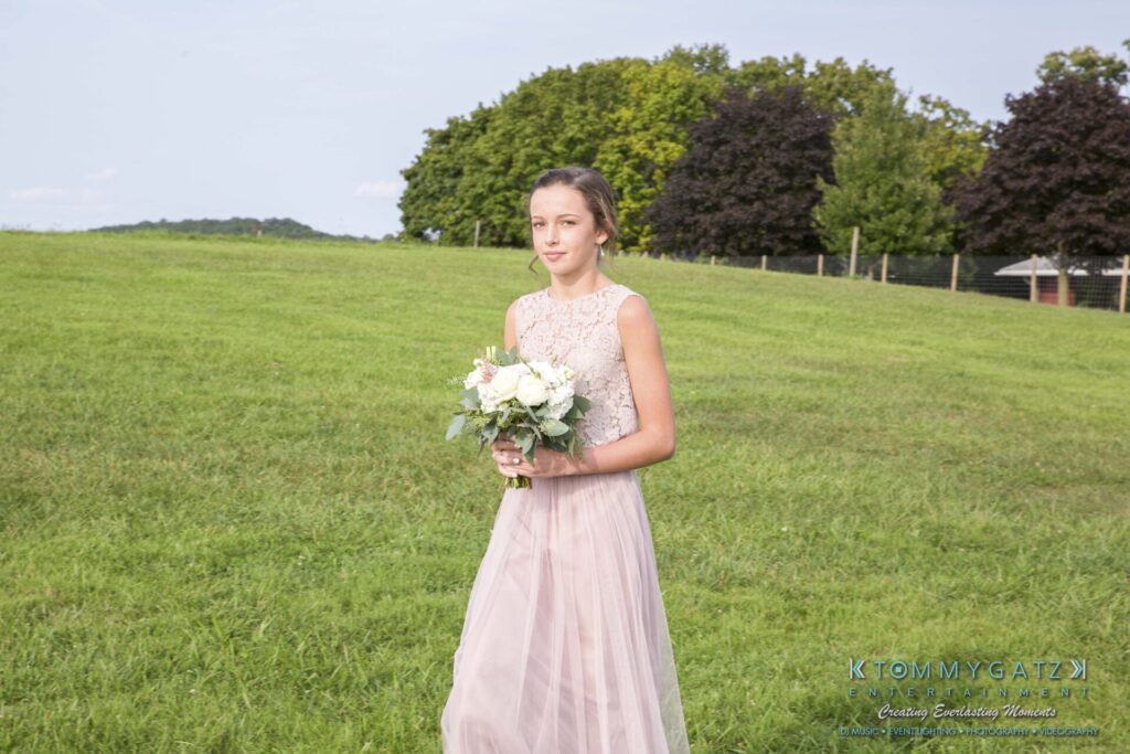 Jennifer-Rudi-Highlights-Low-res-9 - TOMMY GATZ ENTERTAINMENT A young woman in a pink lace dress holds a bouquet while walking across a grassy field. Trees are in the background.