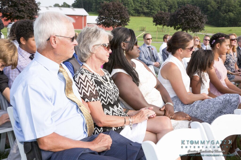 JenniferRudiWeddingCeremonyPhotos-100 - TOMMY GATZ ENTERTAINMENT People seated in rows outdoors during a daytime event, with trees and a red building in the background.