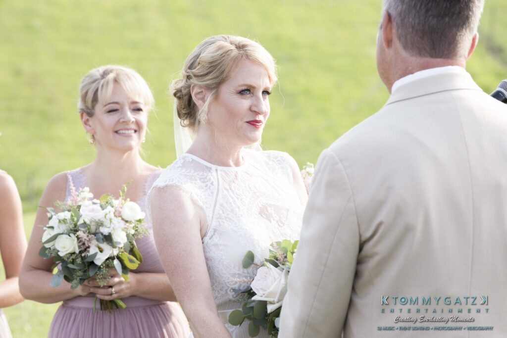 JenniferRudiWeddingCeremonyPhotos-108 - TOMMY GATZ ENTERTAINMENT A bride and groom exchange vows outdoors with a bridesmaid holding a bouquet nearby.