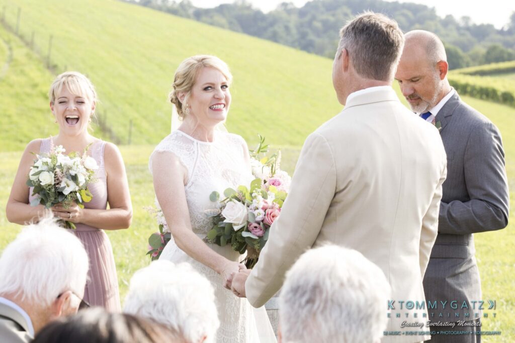 JenniferRudiWeddingCeremonyPhotos-110-1 - TOMMY GATZ ENTERTAINMENT A bride and groom stand facing each other during an outdoor wedding ceremony. The bride holds a bouquet. Bridesmaid and officiant are nearby. The backdrop features a green hillside.