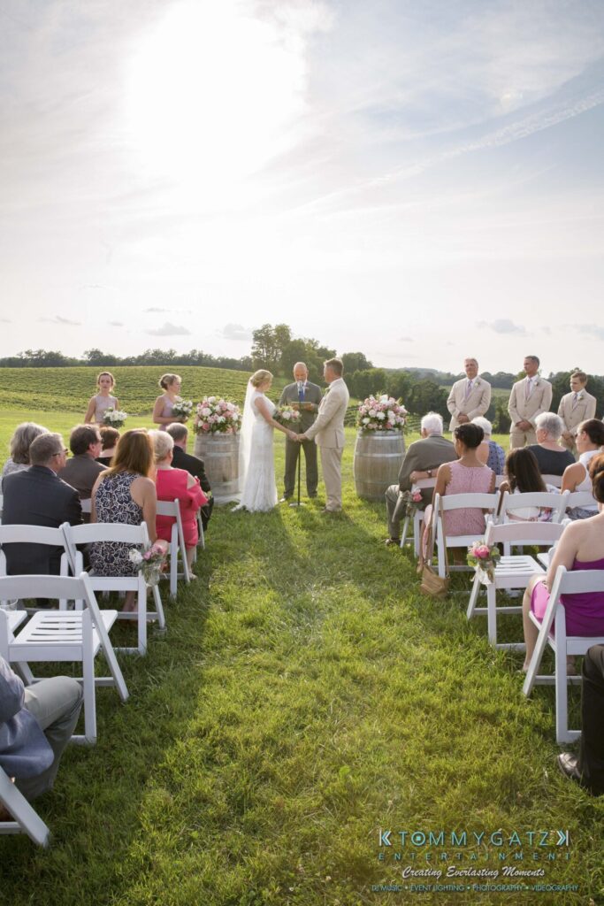 JenniferRudiWeddingCeremonyPhotos-111-1 - TOMMY GATZ ENTERTAINMENT Outdoor wedding ceremony with a couple exchanging vows under bright sunlight. Guests are seated in rows of white chairs on a grassy field.