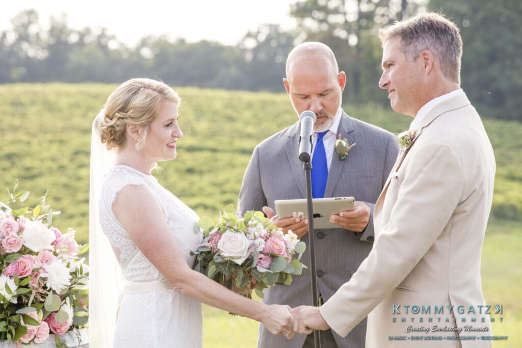 JenniferRudiWeddingCeremonyPhotos-114-1 - TOMMY GATZ ENTERTAINMENT A couple exchanges vows in an outdoor wedding ceremony with an officiant. The bride holds a bouquet of pink and white flowers.