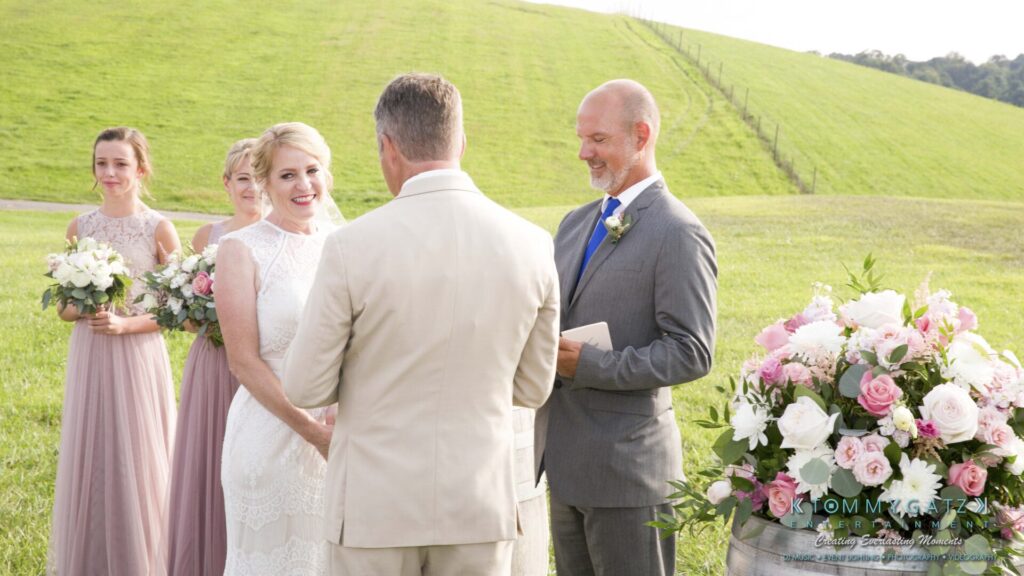 JenniferRudiWeddingCeremonyPhotos-117-1 - TOMMY GATZ ENTERTAINMENT A couple is getting married outdoors. The bride and groom face each other. A man in a suit is officiating. Bridesmaids stand with bouquets. A barrel with flowers is nearby.