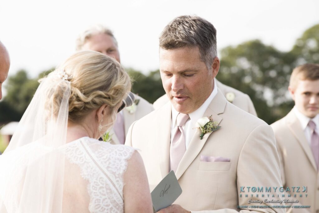JenniferRudiWeddingCeremonyPhotos-118 - TOMMY GATZ ENTERTAINMENT A couple exchanges vows outdoors, dressed in light-colored wedding attire. Bridesmaids and groomsmen stand in the background.