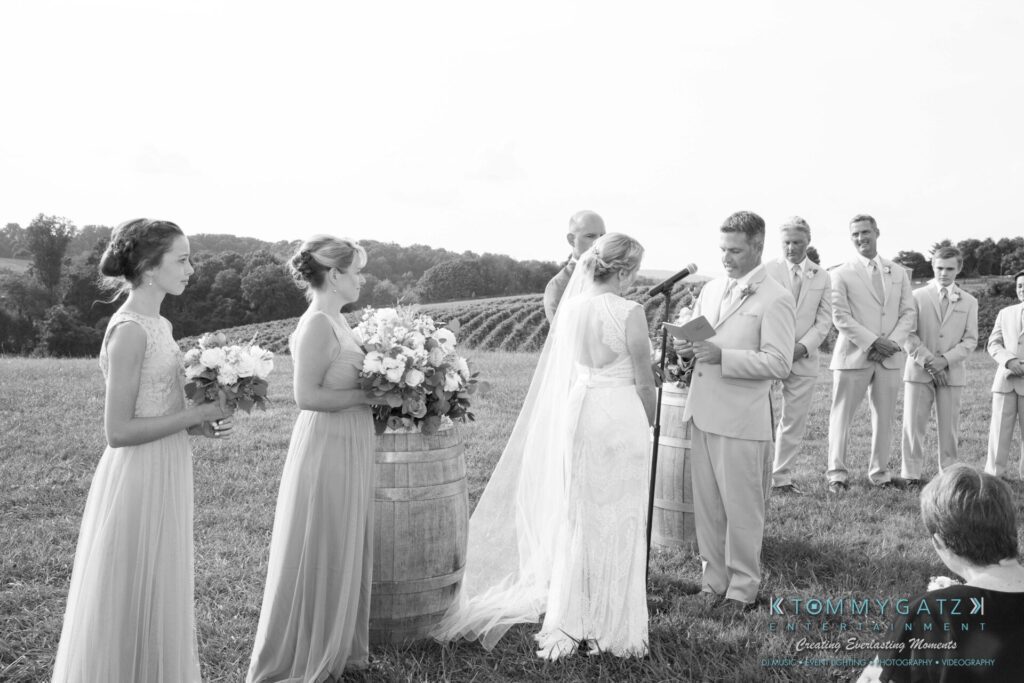 JenniferRudiWeddingCeremonyPhotos-120 - TOMMY GATZ ENTERTAINMENT A couple is getting married outdoors in a vineyard. The groom reads vows while the bride stands beside him. Bridesmaids and groomsmen are present.