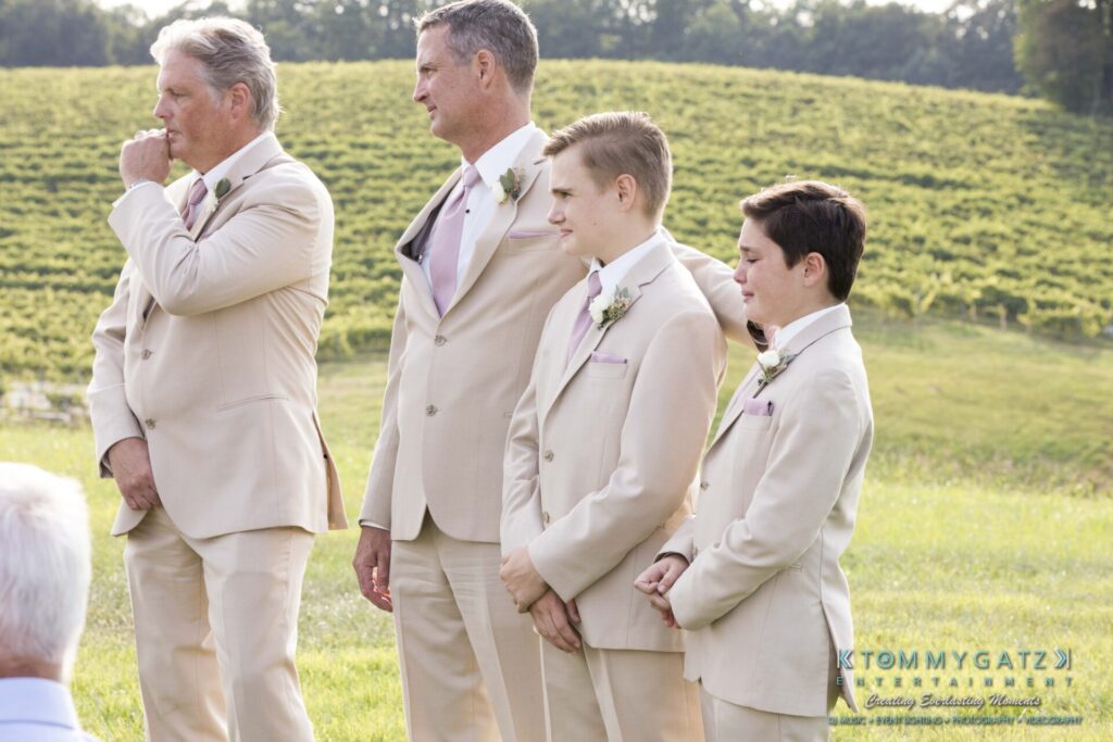 JenniferRudiWeddingCeremonyPhotos-126-1 - TOMMY GATZ ENTERTAINMENT Four men in beige suits stand outdoors on grass with a vineyard in the background.