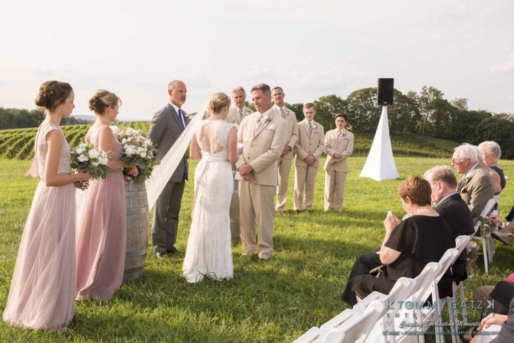 JenniferRudiWeddingCeremonyPhotos-142-1 - TOMMY GATZ ENTERTAINMENT Outdoor wedding ceremony in a field with a bridal party standing and guests seated. The bride and groom face each other, surrounded by a vineyard and greenery.