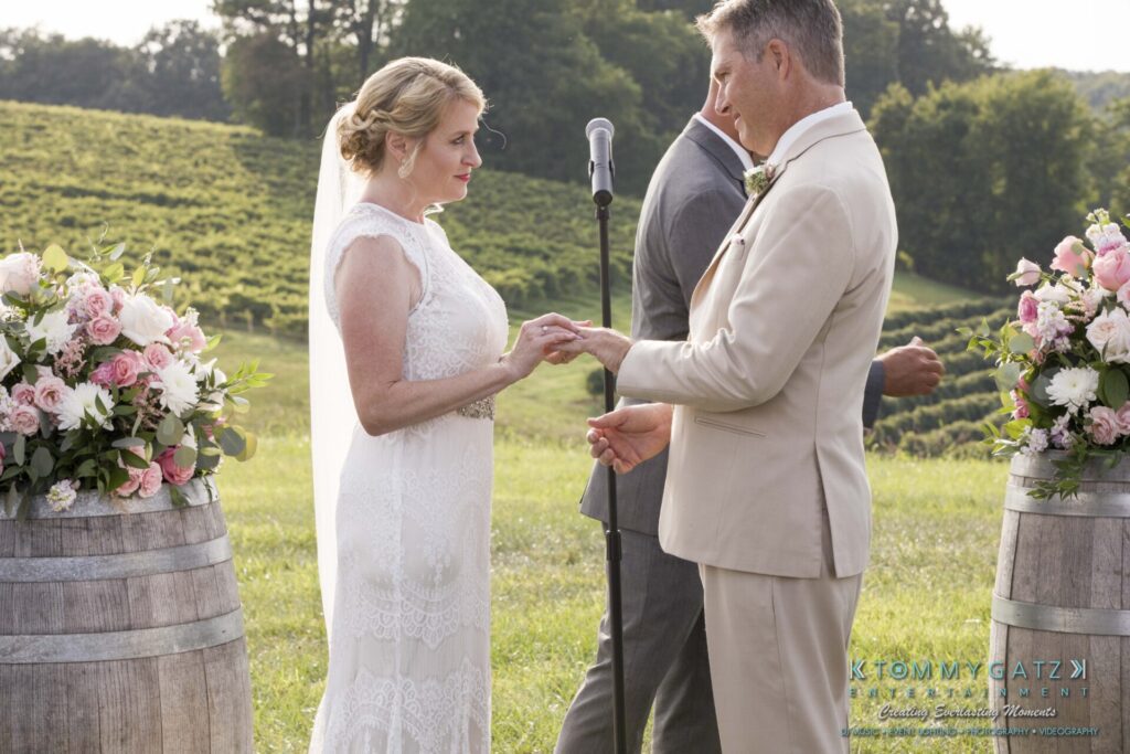 JenniferRudiWeddingCeremonyPhotos-150-1 - TOMMY GATZ ENTERTAINMENT A couple exchanges rings in an outdoor wedding ceremony, flanked by flower arrangements on wooden barrels with a vineyard in the background.