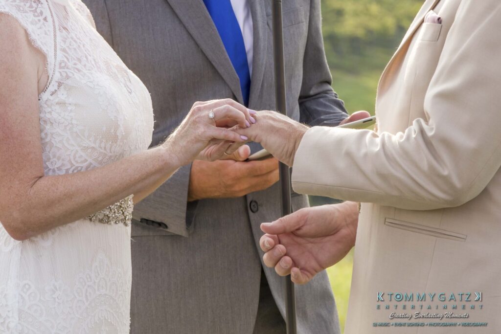 JenniferRudiWeddingCeremonyPhotos-152-1 - TOMMY GATZ ENTERTAINMENT A bride and groom exchange rings during a wedding ceremony outdoors, with a person holding a book in the background.