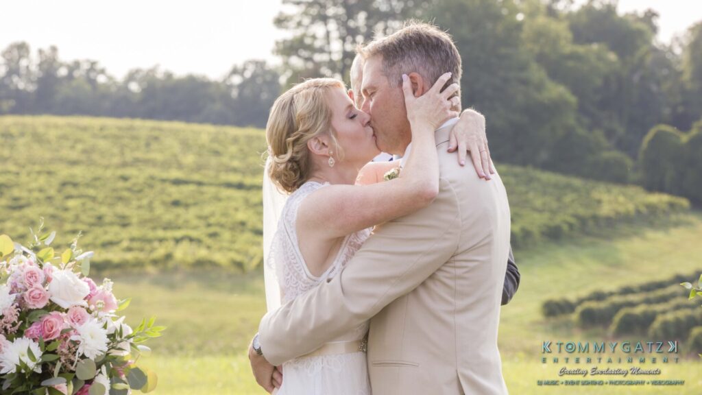 JenniferRudiWeddingCeremonyPhotos-160 - TOMMY GATZ ENTERTAINMENT A couple in wedding attire shares a kiss outdoors near a bouquet and green landscape.
