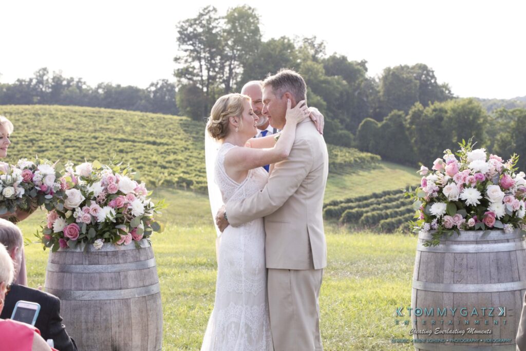 JenniferRudiWeddingCeremonyPhotos-164-1 - TOMMY GATZ ENTERTAINMENT A couple embraces during an outdoor wedding ceremony, surrounded by flowers and wooden barrels, with a vineyard and trees in the background.