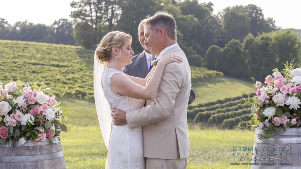 JenniferRudiWeddingCeremonyPhotos-165-1 - TOMMY GATZ ENTERTAINMENT A bride and groom hold each other during an outdoor wedding ceremony, with floral arrangements in the foreground and a vineyard in the background.