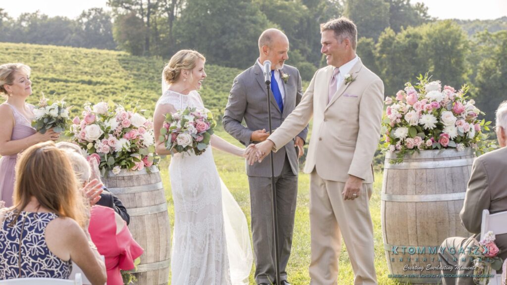 JenniferRudiWeddingCeremonyPhotos-169 - TOMMY GATZ ENTERTAINMENT A wedding ceremony outdoors with a bride in a lace dress and a groom in a light suit shaking hands with a person officiating. Guests are seated, and barrels with flowers are in the background.