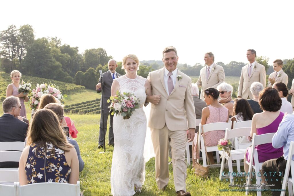 JenniferRudiWeddingCeremonyPhotos-171-1 - TOMMY GATZ ENTERTAINMENT A couple walks down the aisle outdoors after a wedding ceremony, surrounded by guests seated on white chairs.