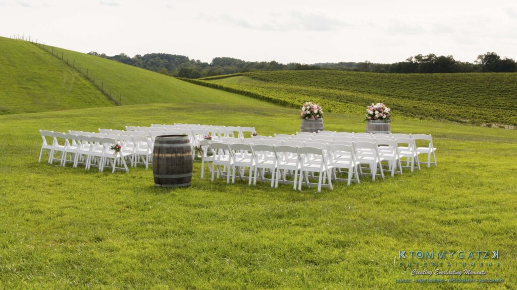 JenniferRudiWeddingCeremonyPhotos-52 - TOMMY GATZ ENTERTAINMENT Outdoor wedding setup with rows of white chairs facing a barrel and floral arrangements, on a grassy hillside.