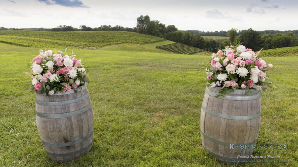 JenniferRudiWeddingCeremonyPhotos-55 - TOMMY GATZ ENTERTAINMENT Two wooden barrels topped with pink and white floral arrangements stand in a grassy field with vineyards and trees in the background.
