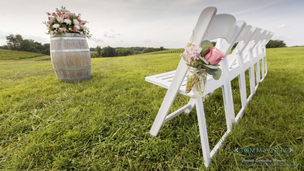 JenniferRudiWeddingCeremonyPhotos-56 - TOMMY GATZ ENTERTAINMENT White chairs arranged on grass with flowers in jars, a wooden barrel with a floral arrangement nearby, and rolling fields in the background.