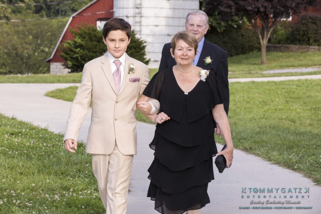JenniferRudiWeddingCeremonyPhotos-62 - TOMMY GATZ ENTERTAINMENT A young boy in a beige suit escorts an elderly woman in a black dress and an older man in a dark suit outside.