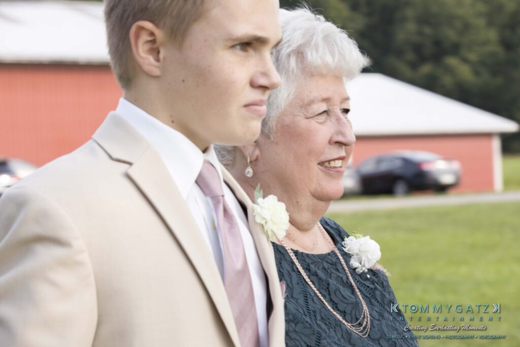 JenniferRudiWeddingCeremonyPhotos-69 - TOMMY GATZ ENTERTAINMENT A young man in a beige suit and an older woman in a teal dress, both wearing floral boutonnieres, walk outdoors near a red building.