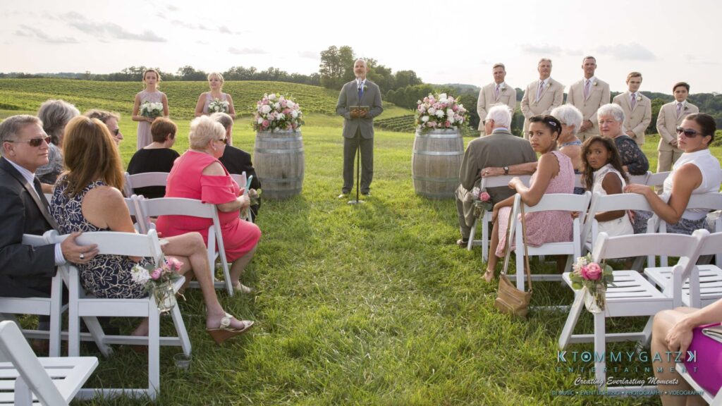 JenniferRudiWeddingCeremonyPhotos-90 - TOMMY GATZ ENTERTAINMENT Outdoor wedding ceremony with guests seated on white chairs, facing an officiant and wedding party. Floral arrangements are on barrels. The setting is a grassy field under a cloudy sky.