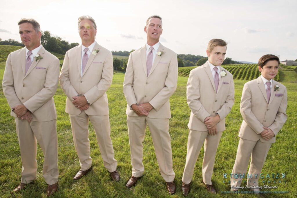 JenniferRudiWeddingCeremonyPhotos-93 - TOMMY GATZ ENTERTAINMENT Five men in beige suits stand on a grassy field with vineyards in the background.