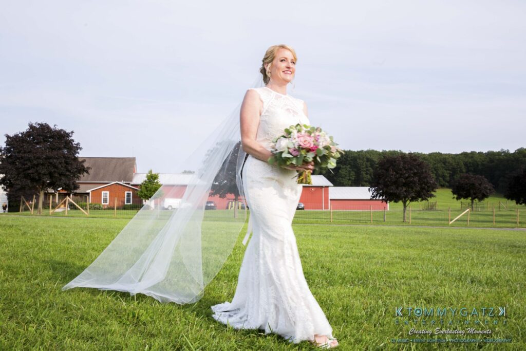 JenniferRudiWeddingCeremonyPhotos-95 - TOMMY GATZ ENTERTAINMENT Bride in a white dress holding a bouquet, walking on a grassy field with barns and trees in the background.