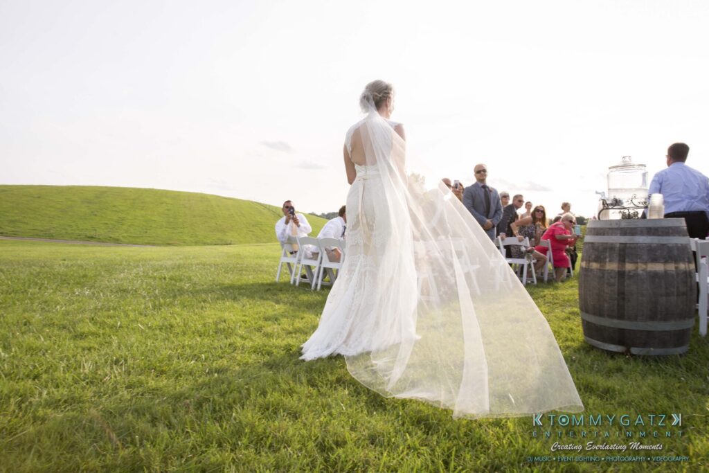 JenniferRudiWeddingCeremonyPhotos-96 - TOMMY GATZ ENTERTAINMENT A bride in a flowing gown walks across a grassy field toward seated guests at an outdoor wedding. A barrel and drink dispenser are visible on the right side.